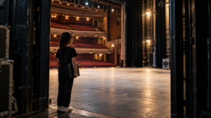 A young performer holding an audition folder stands backstage and looks across an empty professional theatre in Singapore.